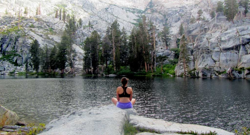 A person sits on a tall rock overlooking a body of water, evergreen trees and mountains in the background.